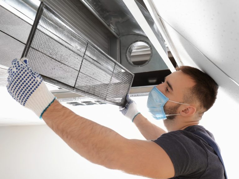 A technician cleaning an air conditioning unit filter while wearing gloves and a mask.